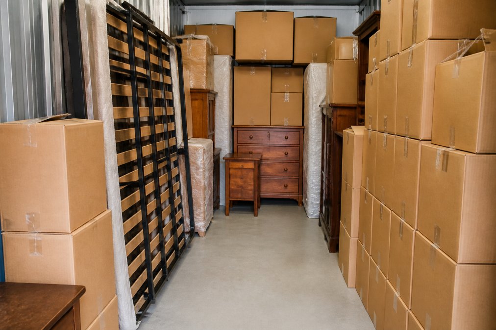 Interior view of a professionally organised storage unit showing neatly stacked moving boxes in uniform rows, a bed frame positioned vertically against one wall, wooden furniture arranged along the back, clear pathways down the centre, and boxes stacked from floor to ceiling in organised columns