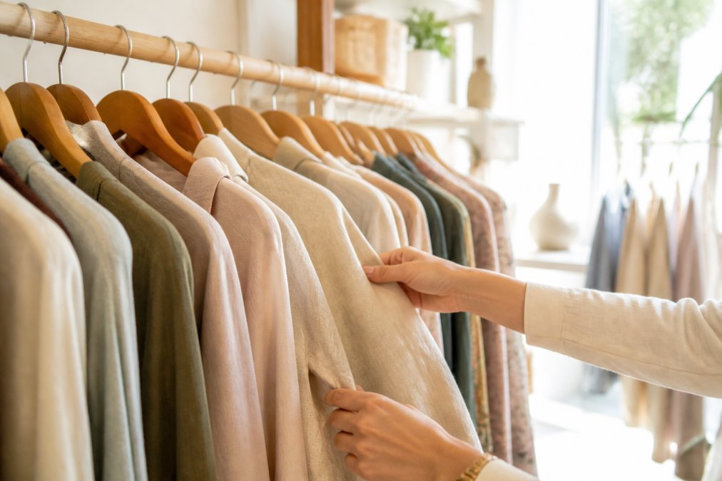 Mani che sfogliano con cura capi di abbigliamento di qualità su un appendiabiti in legno, luce naturale che illumina i tessuti in un ambiente elegante e luminoso