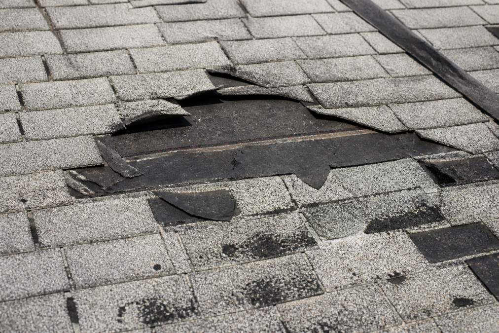 Close-up of roof showing missing shingles, curled and lifted shingle edges, exposed dark underlayment, and scattered granules on light-colored shingles after storm damage
