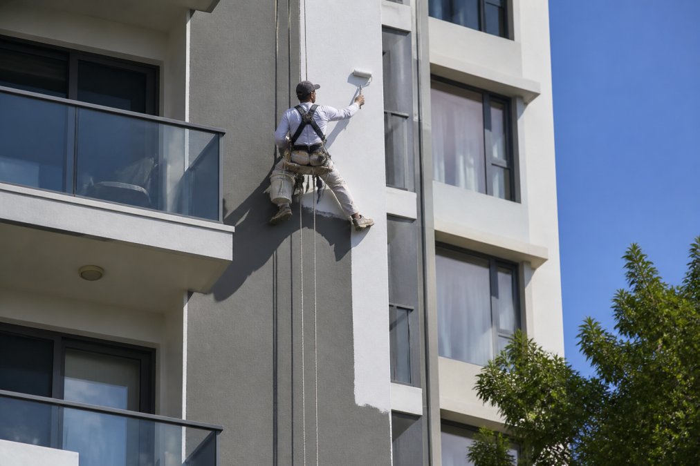 Professional painter in a safety harness applying paint to the exterior facade of a multi-storey residential apartment building on a sunny morning