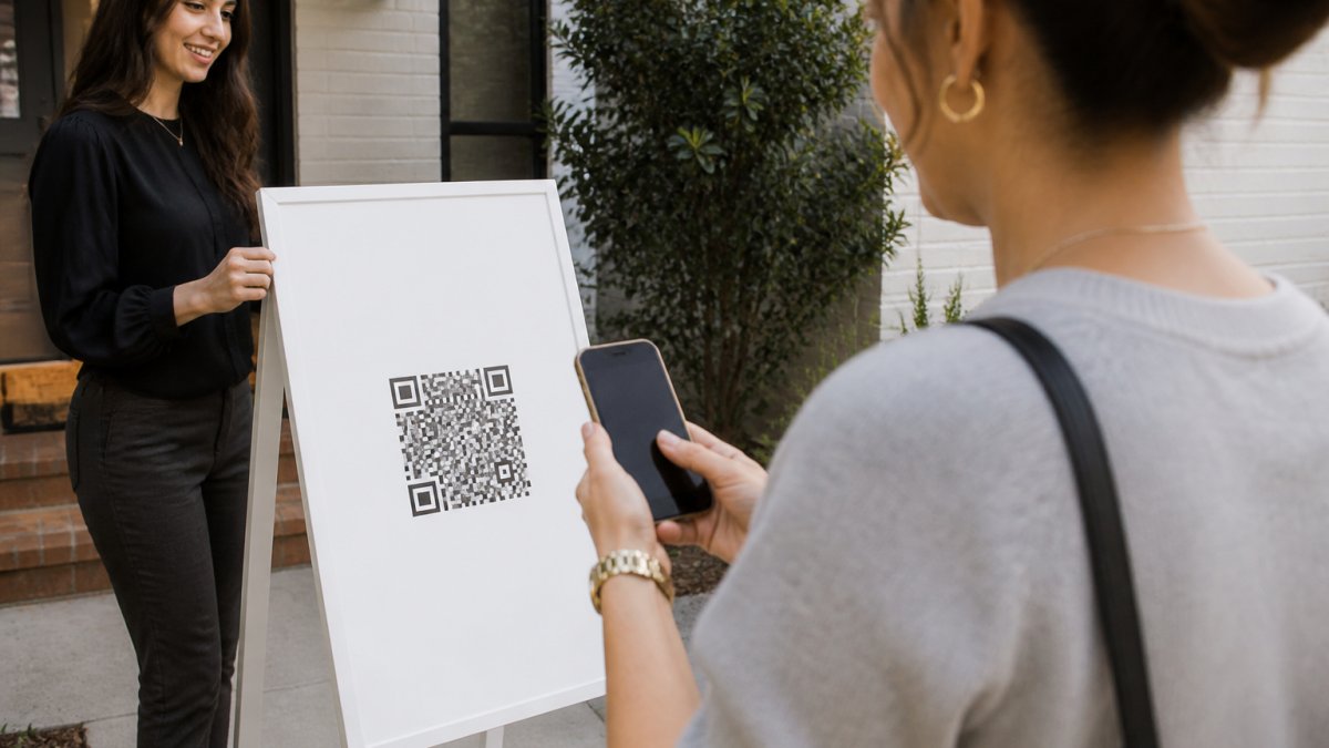 Real estate agent standing outside a home with a branded QR code sign on a white pedestal easel, visitor in casual clothing scanning the QR code with their smartphone