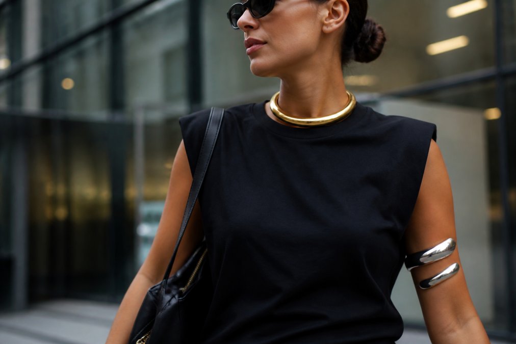A close-up of a woman wearing a bold gold torque choker and sculptural silver arm cuff, layered over a minimalist black top, with soft natural lighting highlighting the metal textures