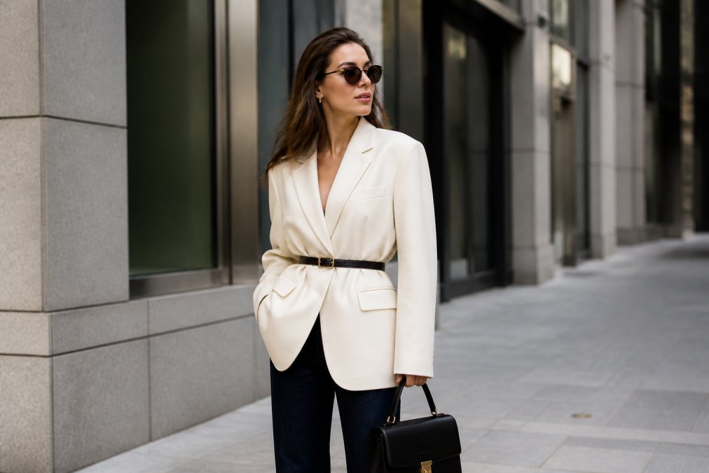 Professional woman wearing oversized tailored cream blazer with cinched waist belt, paired with reliable dark denim and structured leather top-handle bag, standing in urban setting with modern architecture backdrop