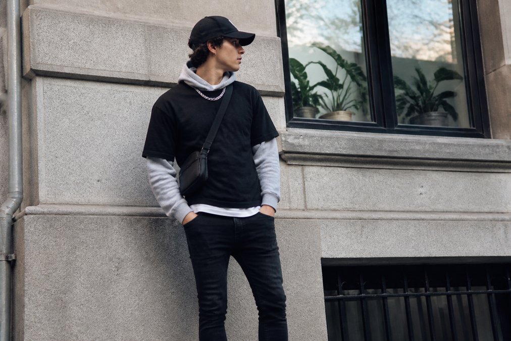 Young person wearing layered streetwear outfit with visible black Signature Cap, chunky silver chain necklace, crossbody bag, and fitted black jeans, standing in urban setting with natural daylight, demonstrating balanced proportions and contrast between fitted and relaxed pieces