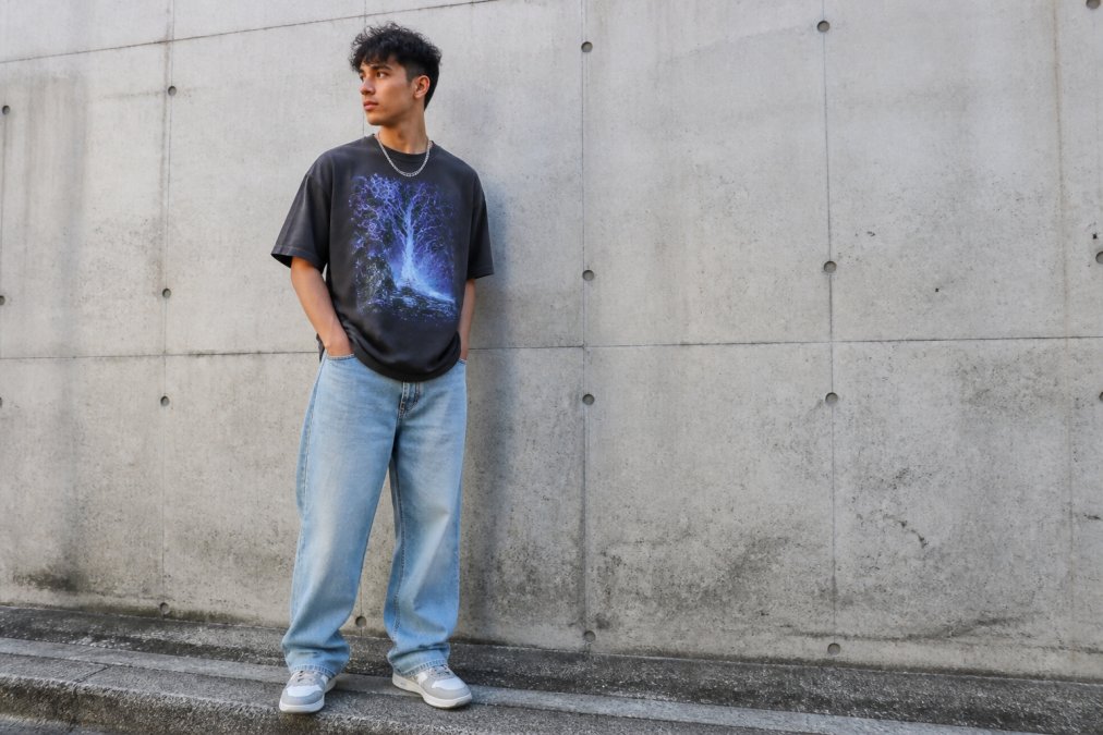 A young adult wearing an oversized graphic tee with relaxed jeans and sneakers, standing in an urban setting with a concrete wall and natural daylight, showing how the drop-shoulder fit drapes naturally