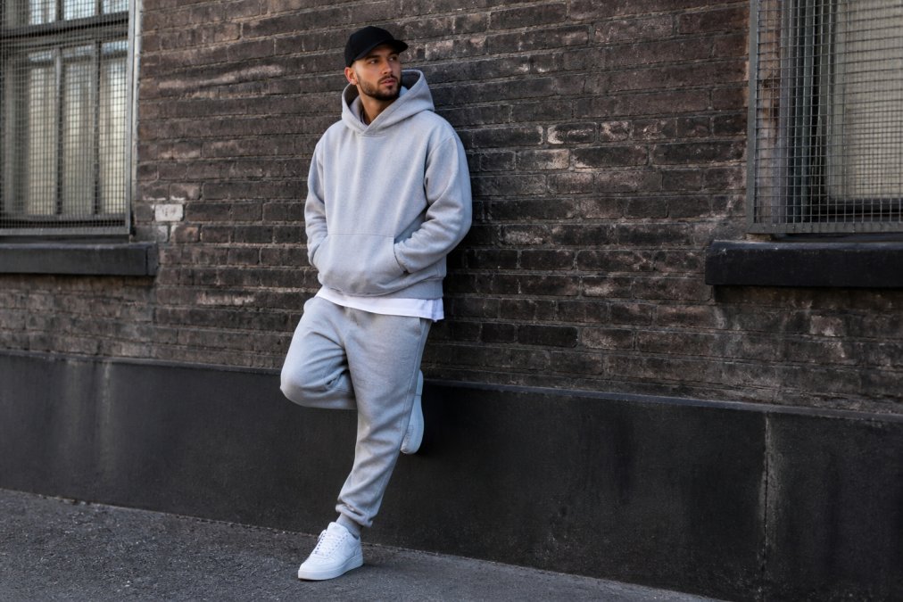 Person i oversized hoodie og joggingbukser med hvide sneakers, afslappet stående positur foran en urban murstensbaggrund, der demonstrerer layering-teknik med synlige teksturforskelle