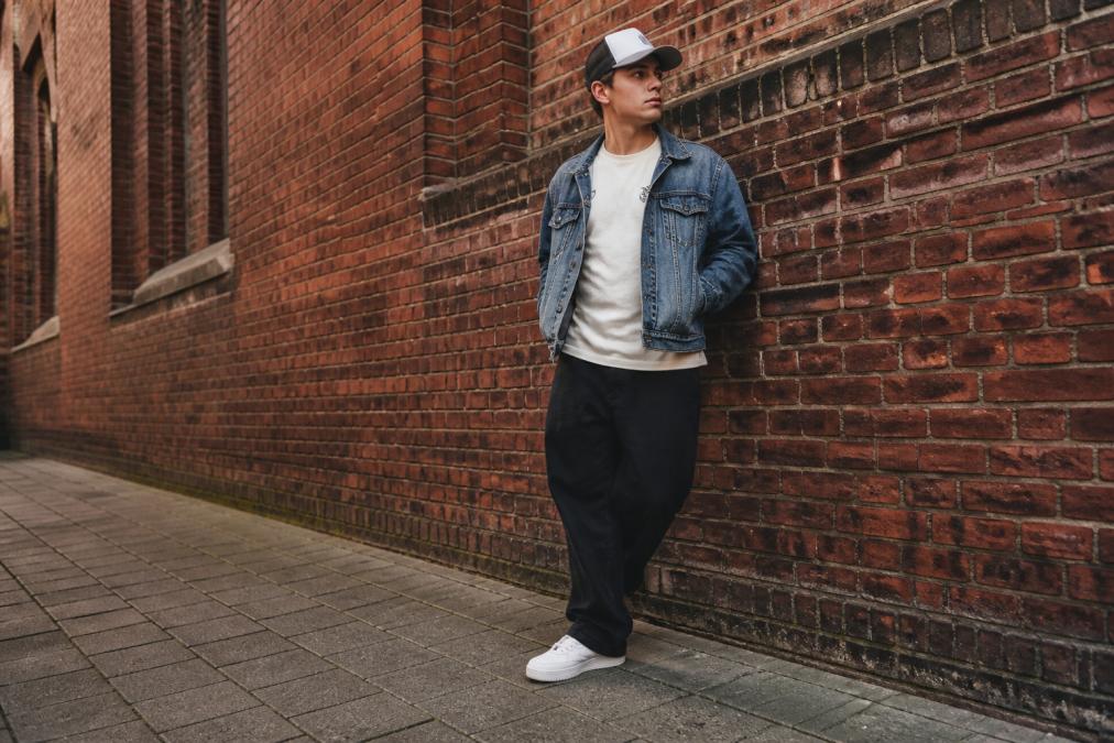 Nærbillede af streetwear-detaljer på en person: grafisk T-shirt med subtilt logo, vintage-denimjakke, truckercap og hvide sneakers, stående i et bymiljø med en baggrund af mursten