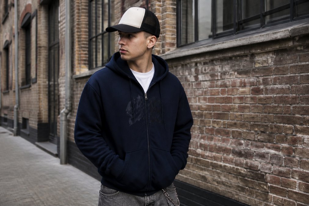 Ung person bär en oversized marinblå hoodie med subtilt grafiskt tryck, truckerkeps och minimalistisk t-shirt i en urban miljö med tegelväggar och diffust dagsljus