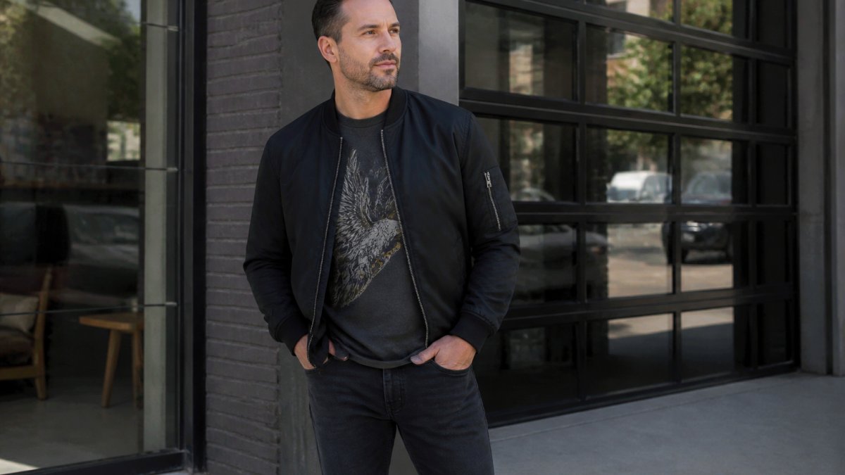 Man in his 30s wearing well-fitted graphic tee layered under casual bomber jacket and dark jeans, standing confidently in natural light with relaxed posture