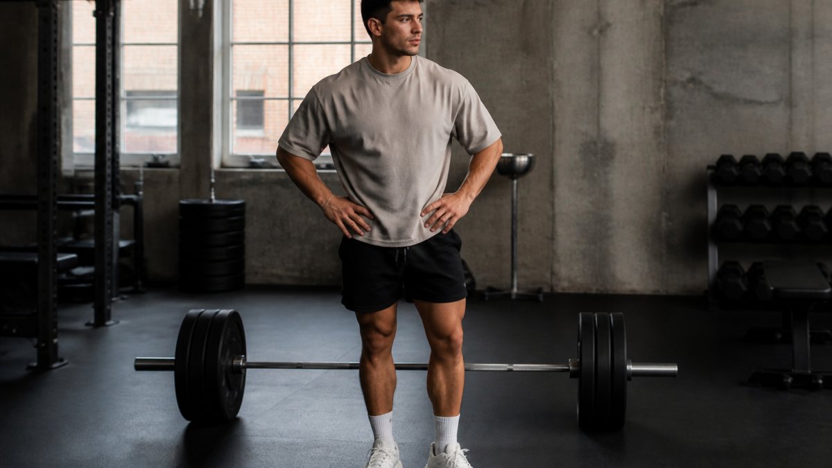 A man in his late twenties wearing an oversized moisture-wicking t-shirt, tapered training shorts, and clean sneakers, standing in a dim industrial gym between sets with a loaded barbell behind him