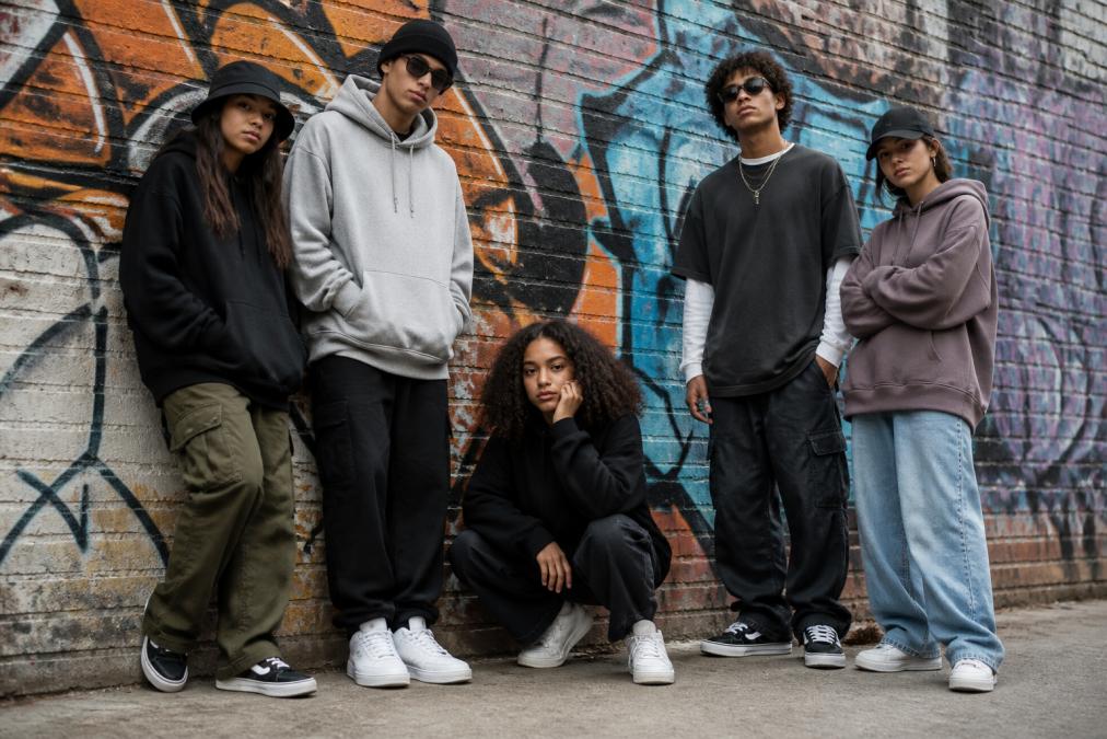 Jongeren in trendy streetwear outfit met oversized hoodies, graphic t-shirts en sneakers in een stedelijke omgeving, staand tegen een bakstenen muur met graffiti