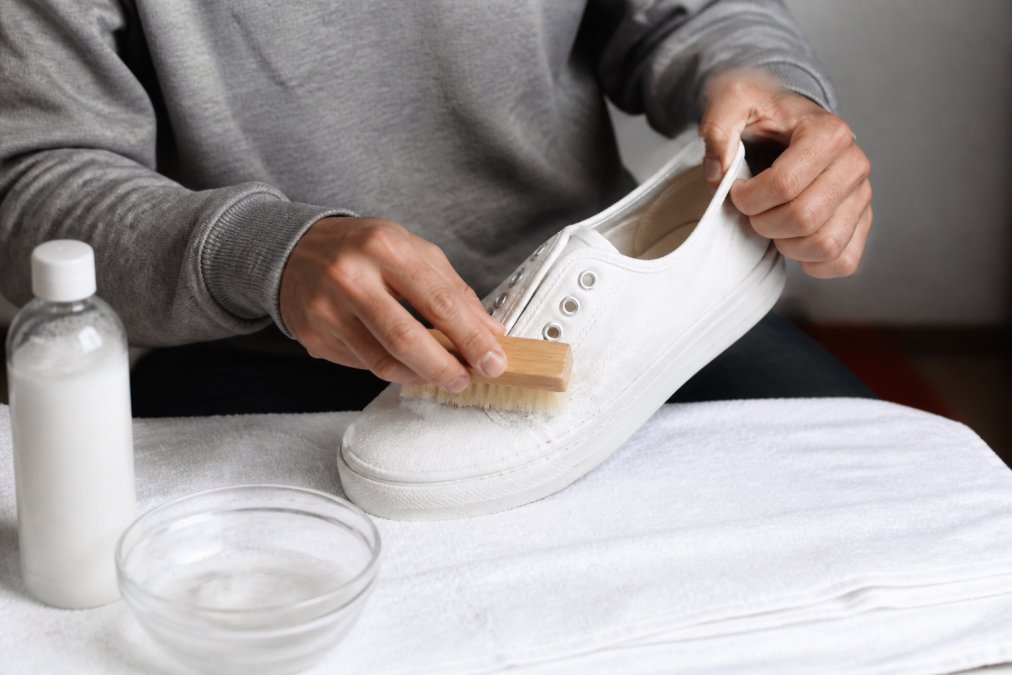 Iemand die voorzichtig een witte canvas sneaker schoonmaakt met een zachte borstel en mild reinigingsvloeistof, met de sneaker op een schone witte handdoek