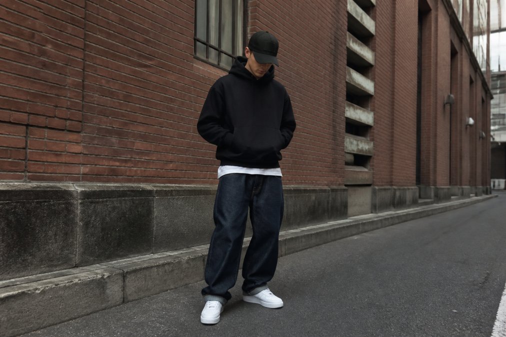 Ung mand i oversized sort hoodie og baggy jeans med hvide sneakers, stående i urbane omgivelser foran en murstensvæg, der demonstrerer, hvordan layering og balance skaber et modent streetwear-look