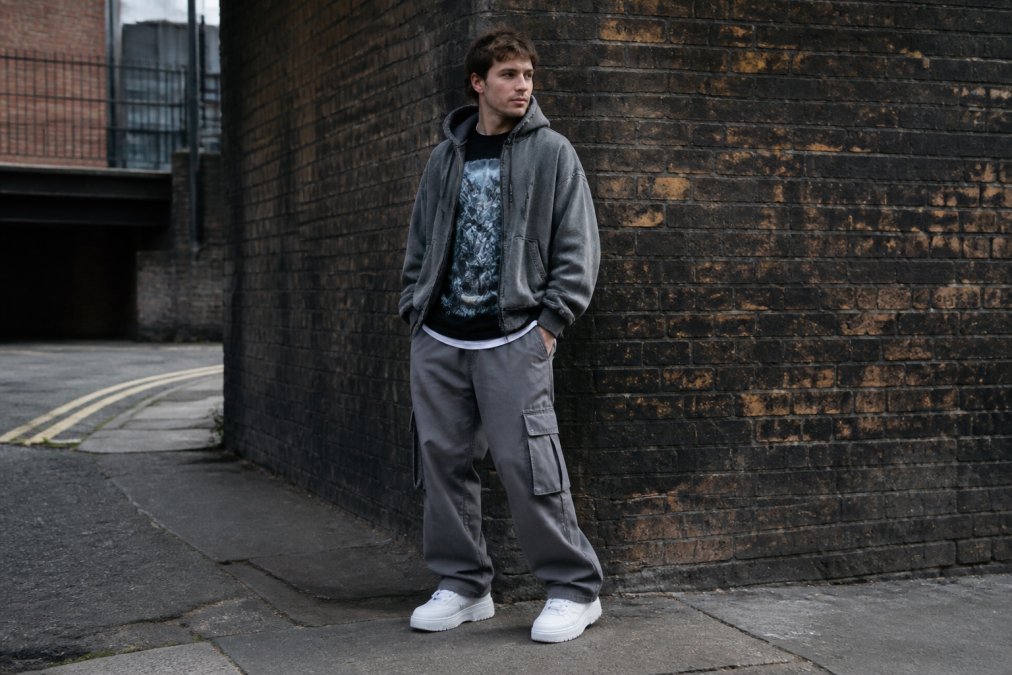 Person in a complete streetwear outfit with an oversized hoodie layered over a graphic tee, baggy cargo pants, and chunky white sneakers, standing in an urban street setting with a brick wall in the background