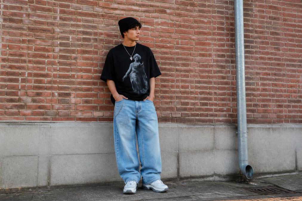 Full photo of person in a complete streetwear look with oversized graphic tee, baggy jeans, chunky sneakers, and streetwear accessories, standing against a brick wall or urban street background with natural city lighting