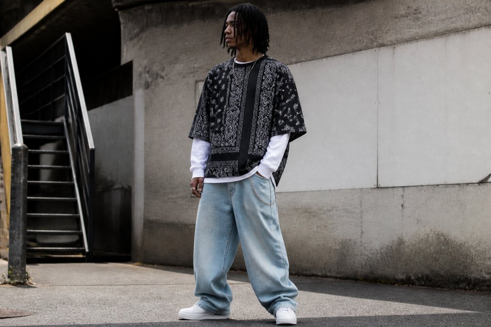 Young person wearing oversized baggy jeans, an oversized bandana-patterned t-shirt, and layered streetwear pieces standing in an urban environment with natural daylight, demonstrating relaxed proportions and comfortable silhouette
