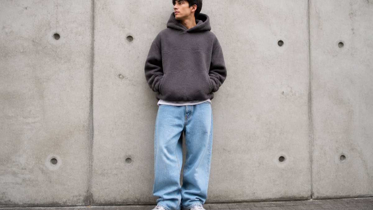A young adult wearing an oversized boxy fleece hoodie with baggy jeans and chunky sneakers, standing against a concrete wall in an urban setting, natural daylight, full-body street style shot