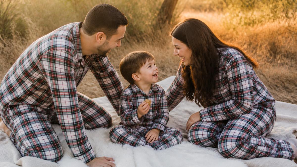A relaxed outdoor family photo scene: parents in coordinating plaid loungewear kneeling on a blanket with a toddler holding a small snack, golden late-afternoon light, everyone laughing naturally