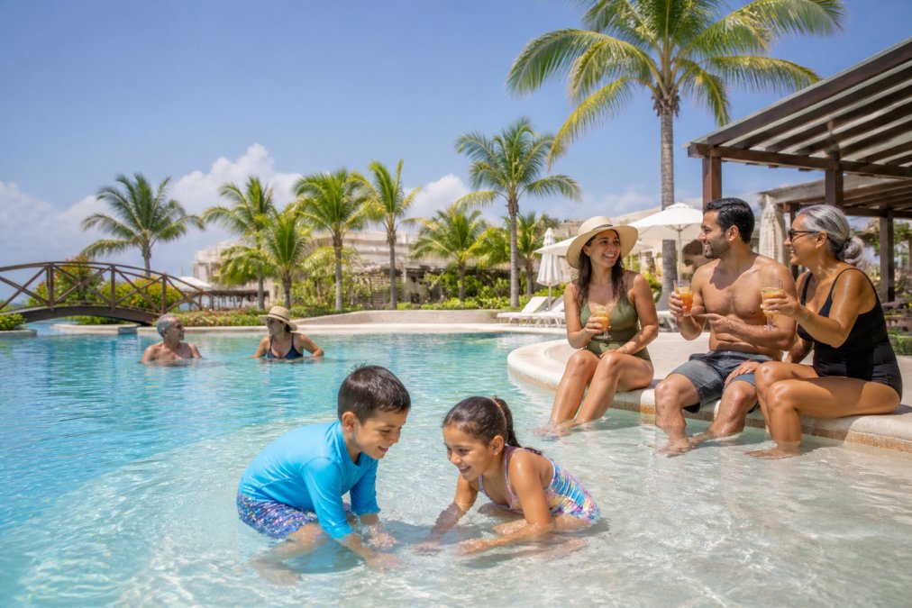 Multi-generational family relaxing together at resort pool, kids playing in shallow end while parents sit nearby with drinks, tropical setting with palm trees and clear sky