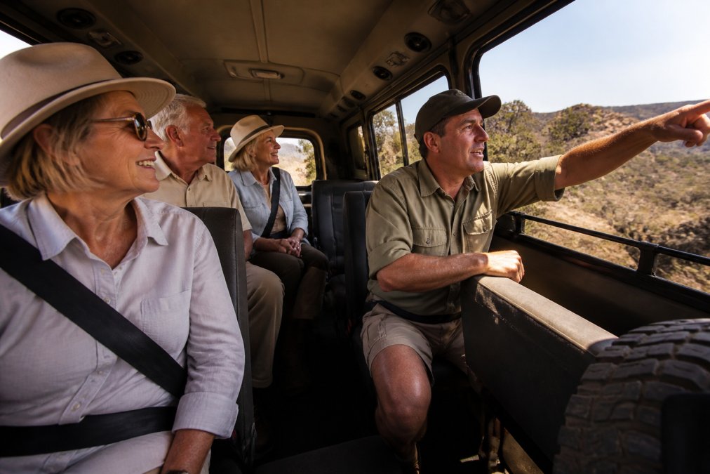 A group of mature travellers sitting together in a comfortable 4WD vehicle, smiling and engaged with an expert guide pointing out scenery through the window