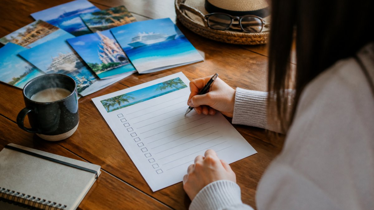 Person sitting at a wooden desk with a printed vacation planning checklist, pen in hand, laptop open nearby, with colorful travel brochures and a steaming cup of coffee visible on the desktop