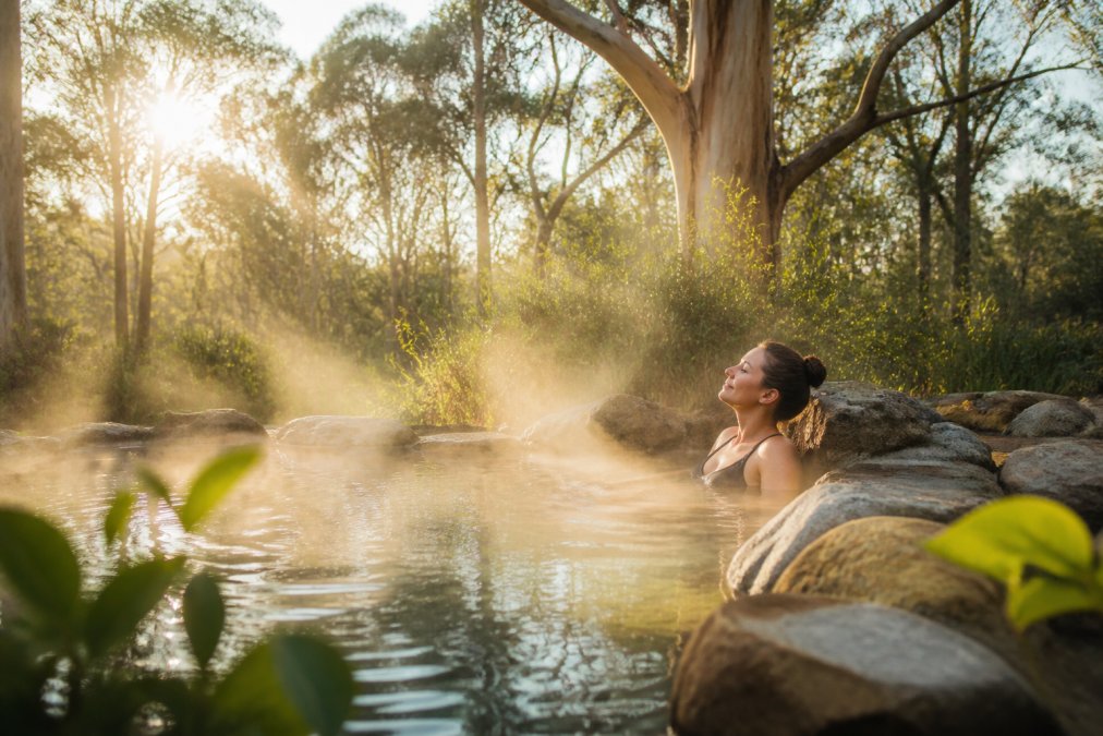 A guest relaxing in a natural thermal pool surrounded by native vegetation and natural stone, with soft morning light filtering through eucalyptus trees, creating a serene sanctuary atmosphere