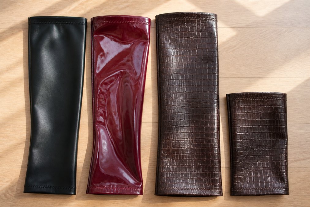 Flat lay of four Bootsy boot sleeves arranged side by side on a light wood surface, black faux leather, deep cherry vinyl, chocolate crocodile thigh-high, and chocolate crocodile mid-calf, shot from directly above in bright natural light