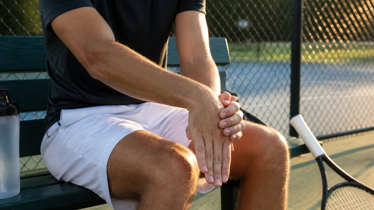 Close-up of an adult tennis player seated on a court bench, using their other hand to gently pull the affected hand into a wrist extension stretch, forearm resting on their thigh, racquet beside them, soft late-afternoon light
