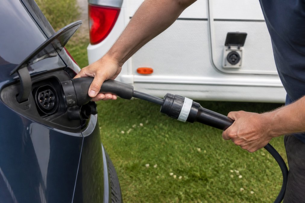 Close-up van handen die een V2L-adapterkabel aansluiten van een elektrische auto op een camper, waarbij de stekker en kabel duidelijk zichtbaar zijn bij daglicht.