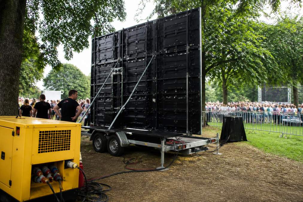 Groot LED-scherm op een aanhangwagen opgesteld op een buitenevenement, met zichtbare stroomkabels en een dieselgenerator op de voorgrond, omgeven door evenementcrew die aansluitingen controleert, gouden namiddaglicht