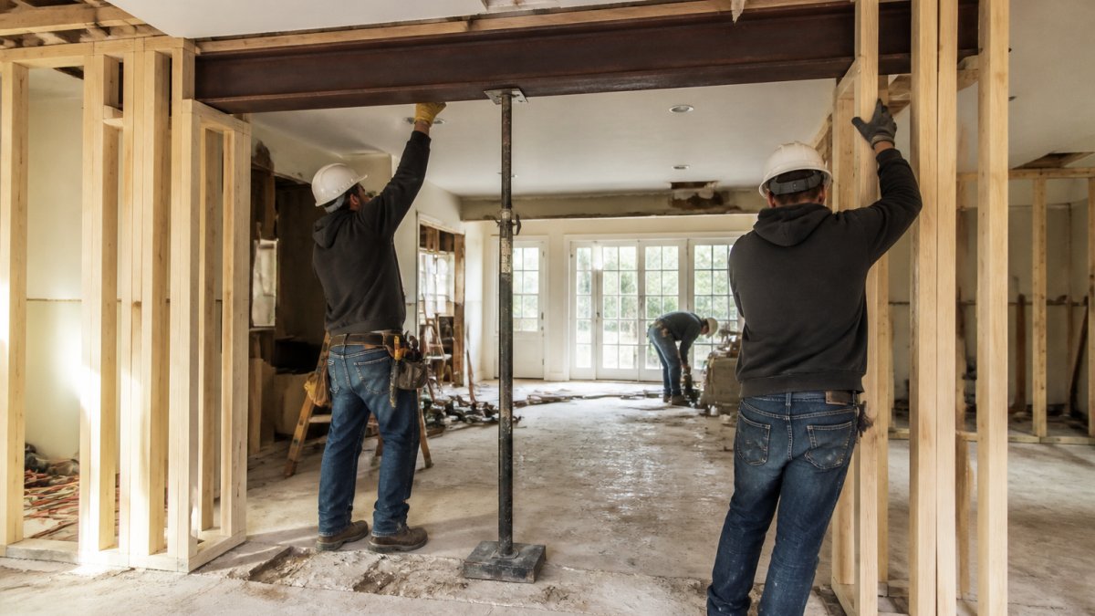 Professional construction team installing a steel support beam in a residential home during wall removal, with temporary wooden support walls visible on both sides, natural daylight streaming through windows