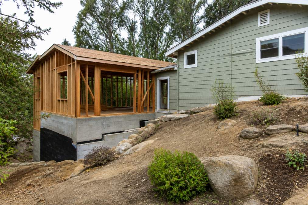 Residential property showing a detached ADU structure in the backyard with visible foundation work, framing, and connection to main house on a sloped lot with natural landscaping