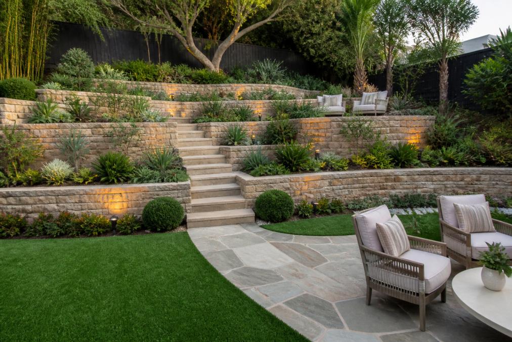 Wide view of a multi-level tiered garden on a sloping residential property showing retaining walls, garden beds, stone pathways and outdoor seating areas