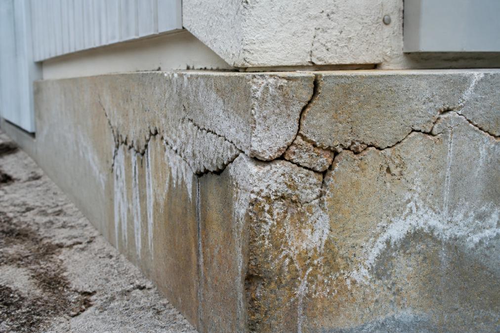 Close-up of concrete foundation showing visible cracks, spalling, and salt-stained mortar on a coastal home exterior wall