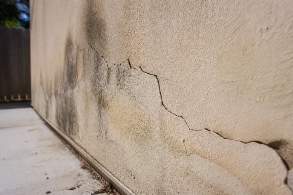 Close-up of exterior stucco wall showing hairline cracks with visible discoloration and moisture staining, sunlight highlighting the deteriorated surface texture and separation at edges