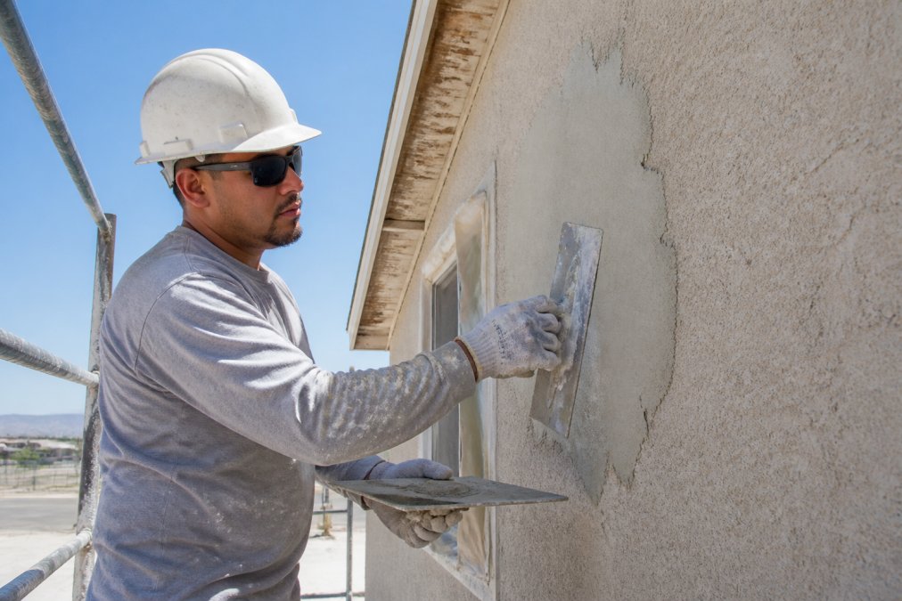 Licensed contractor in work gear and safety equipment applying stucco patch material to exterior wall with professional tools visible, demonstrating proper application technique with natural daylight