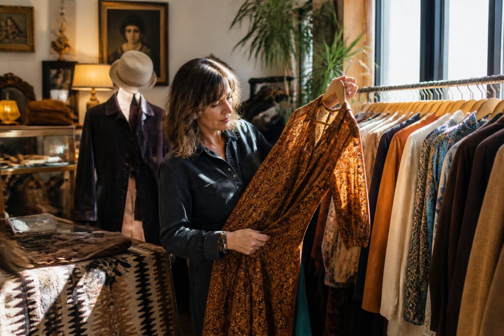 Eine Frau mittleren Alters hält ein handgeprüftes Vintage-Kleidungsstück aus den 1970er Jahren ins Licht, während sie in einem gemütlichen, gut sortierten Second-Hand-Laden steht. Im Hintergrund sind ordentlich aufgereihte Kleiderbügel mit zeitlosen Basics und Retro-Statements zu sehen. Warmes, natürliches Licht fällt durch ein Fenster.