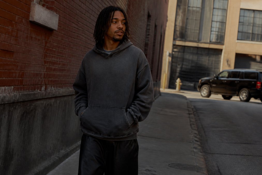 A stylish person in their late 20s wearing a washed, loose-fit hoodie walking through a gritty urban street with brick walls, looking confident and authentic