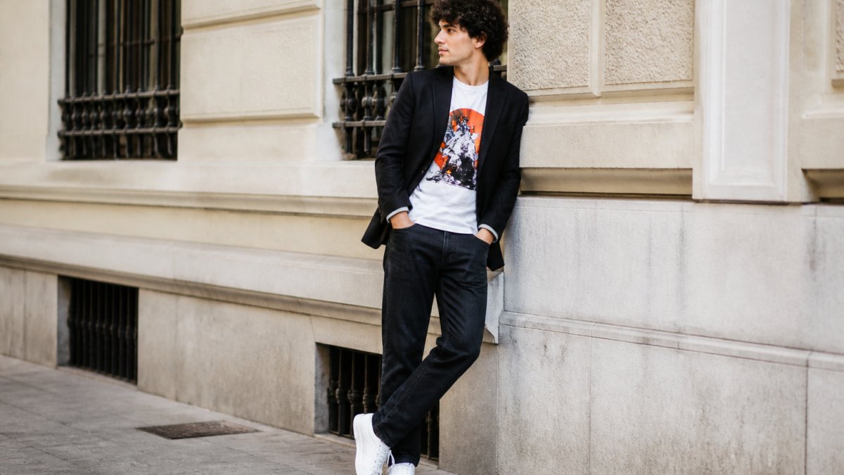 Person in a bold graphic tee and casual blazer standing confidently in natural light with dark jeans and sneakers