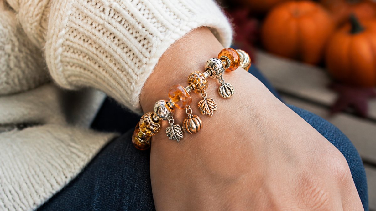 Wrist displaying handmade charm bracelet with autumn-colored beads in warm oranges and golds, paired with a cozy knit sweater sleeve in cream, showing seasonal charms like maple leaves and pumpkins