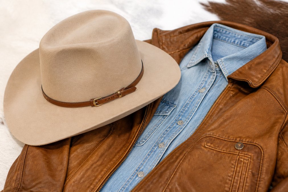 Close-up detail shot showing a classic Stetson hat positioned next to a caramel leather jacket, highlighting how the hat's brim width and structured crown complement the jacket's texture and silhouette