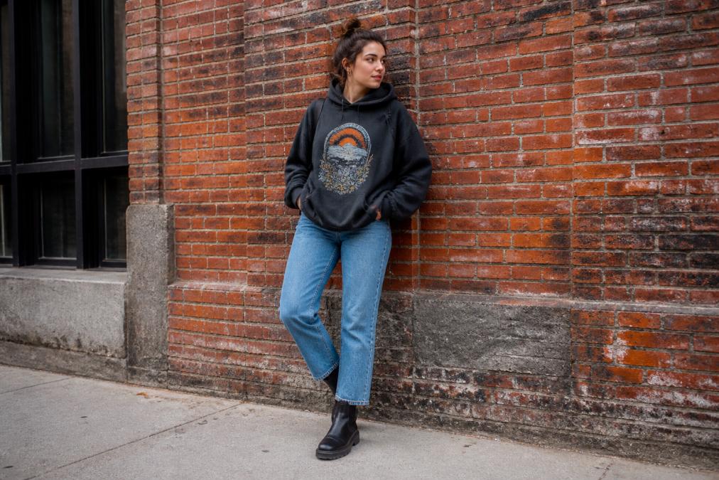 A young adult wearing an oversized graphic hoodie with straight-leg jeans and ankle boots, standing against a brick wall in an urban setting, casual daytime light