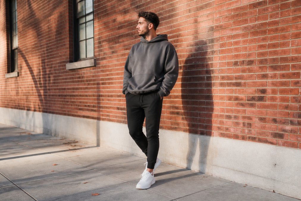 A young adult wearing an oversized hoodie with fitted tapered joggers and clean white sneakers, standing against a brick wall in an urban setting under soft afternoon light
