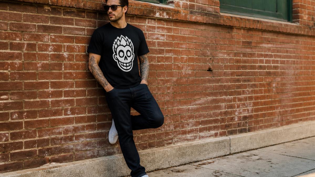 A man wearing a bold graphic t-shirt with dark jeans and white trainers, standing casually outdoors near a brick wall, natural daylight, relaxed posture