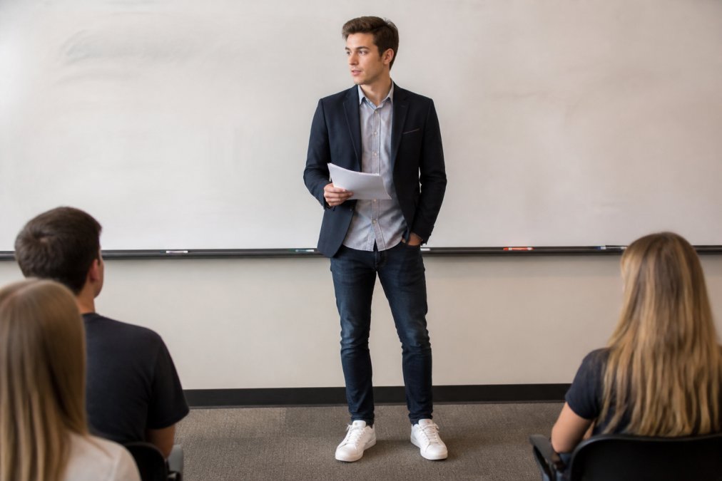 A college student standing confidently in front of a classroom whiteboard, wearing a smart casual blazer over a button-down shirt with dark jeans and clean sneakers, holding presentation notes