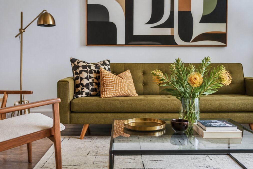 Modern living room with mid-century sofa paired with contemporary abstract art, vintage brass floor lamp, and modern geometric throw pillows arranged on a glass coffee table with books and fresh flowers