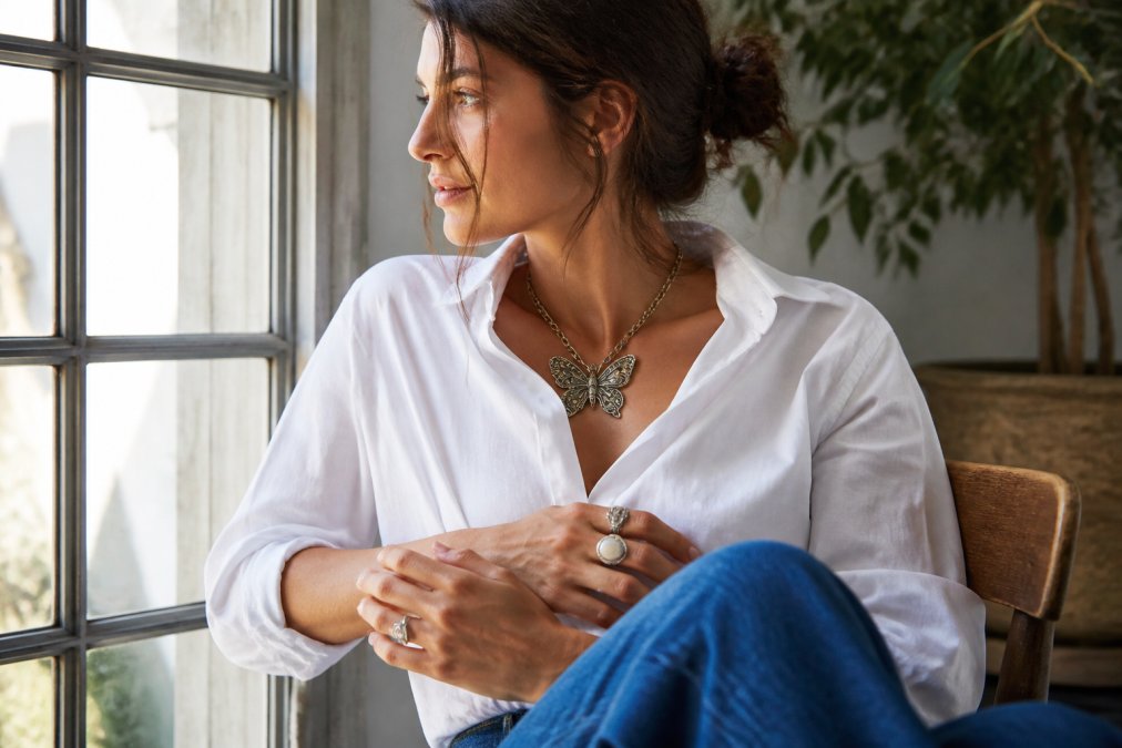 Woman wearing crisp white button-down shirt and jeans, styled with bold vintage butterfly necklace and stacked rings, photographed in natural daylight near a window