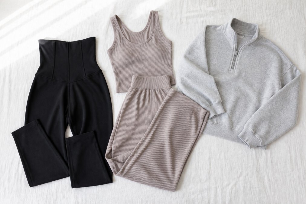 Flat lay of stylish activewear pieces including black corset leggings, a ribbed tank and wide leg pants set in neutral tones, and a heather gray half-zip sweatshirt arranged neatly on a white linen background with soft natural window light