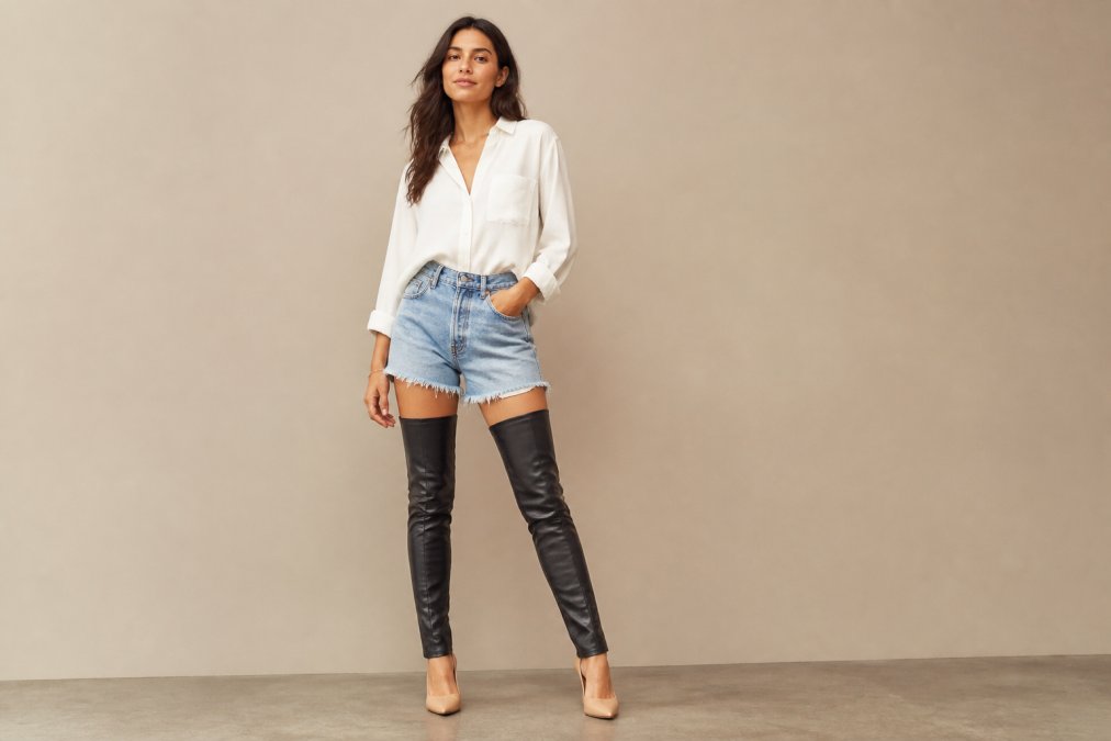 Woman wearing a black thigh-high boot sleeve over a nude heel with a casual outfit, standing confidently against a neutral background