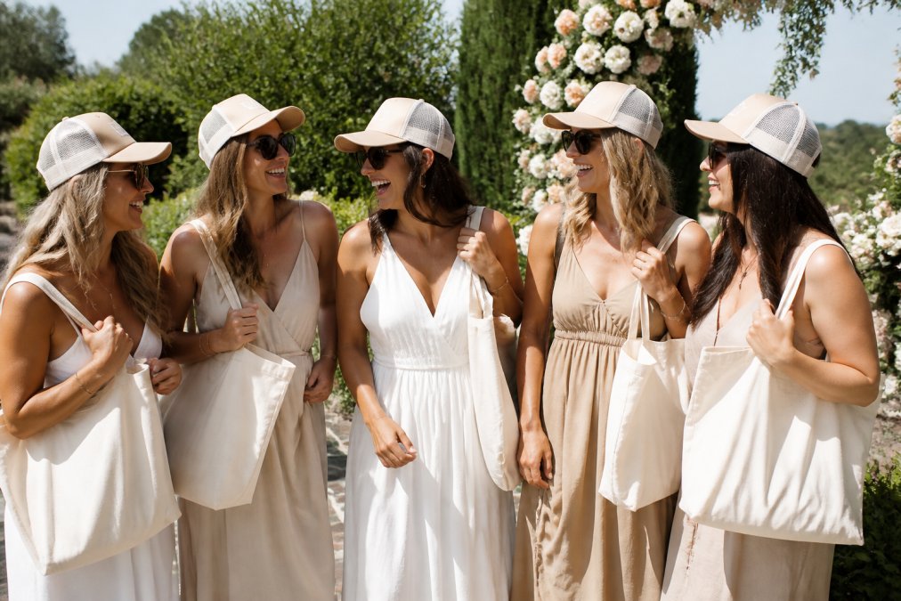 A group of bridesmaids wearing matching trucker hats and holding canvas tote bags, laughing together in a sunny outdoor setting with floral decorations